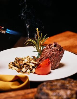A beautifully plated steak with herbs, grilled vegetables, and a charred tomato, enhanced by a culinary torch for a dramatic touch.