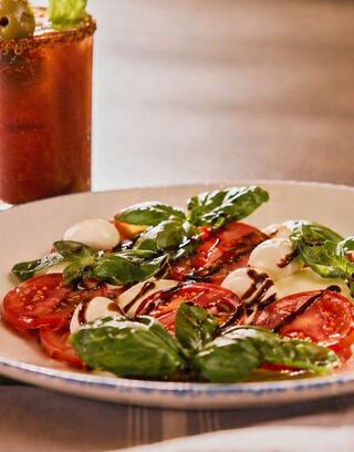 A colorful plate features fresh tomatoes, mozzarella, and basil drizzled with balsamic, alongside a refreshing drink.