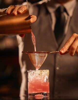 A bartender skillfully pours a vibrant pink cocktail through a strainer into a glass, highlighting craftsmanship and attention to detail.