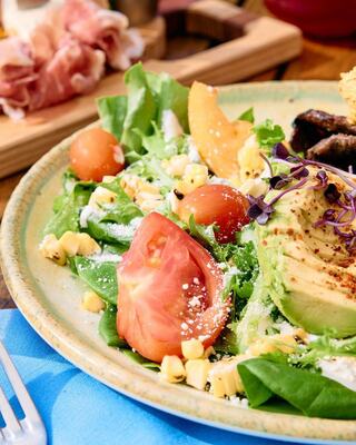 A colorful salad features greens, cherry tomatoes, corn, avocado, and dressing, garnished with microgreens and paired with cured meats in the background.
