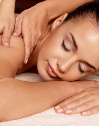 A woman relaxes with her eyes closed, enjoying a soothing massage, as gentle hands work on her shoulders amidst a tranquil setting.