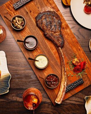 A large, grilled tomahawk steak rests on a wooden board, surrounded by various dipping sauces and garnished with roasted vegetables.