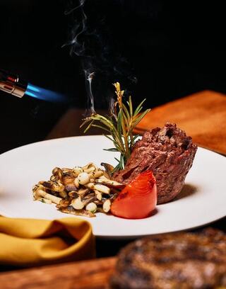 A gourmet dish features a perfectly cooked piece of meat, garnished with herbs, roasted tomato, and sautéed mushrooms, with a chef's torch in action.