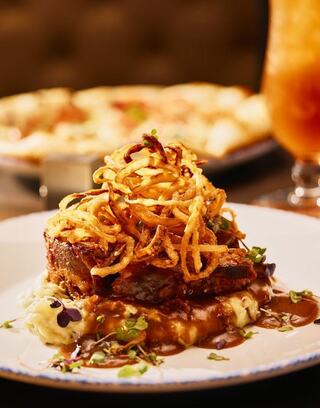 A savory dish features a hearty meat portion topped with crispy fried onions, nestled on creamy mashed potatoes and garnished with greens.