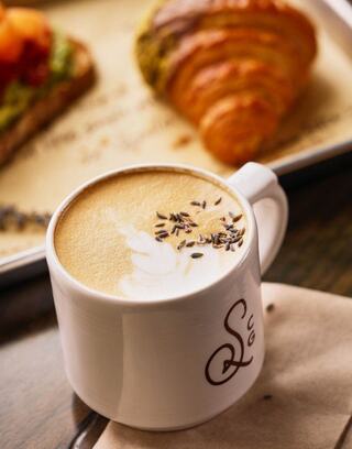 A steaming cup of lavender coffee topped with frothy milk sits beside a golden croissant, creating a cozy café vibe.