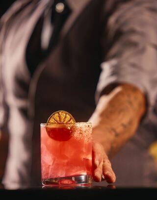A bartender serves a vibrant, garnished cocktail with a slice of dried lime, highlighted by moody lighting and elegant attire.