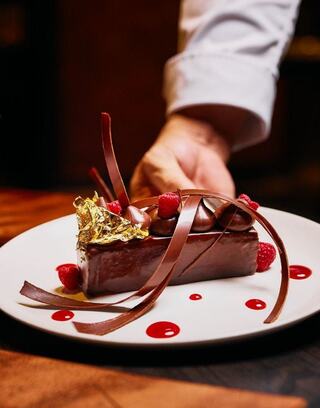 A rich chocolate dessert garnished with raspberries, gold leaf, and elegant chocolate curls, elegantly arranged on a plate.