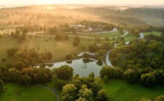 A serene landscape features lush greenery, a winding river, and a tranquil pond, enveloped in soft morning mist and gentle light.