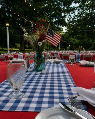 A festive outdoor dining setup features red tables with checkered table runners, flower arrangements, and small American flags.