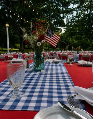 A festive outdoor dining setup features red tables, checkered tablecloths, glassware, and a bouquet with an American flag centerpiece.