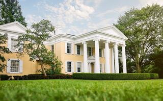 Oglebay Mansion features classic white columns and lush green landscaping under a bright, partly cloudy sky.