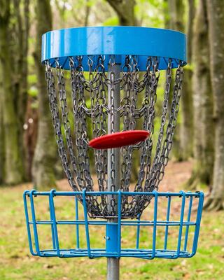 A disc golf basket stands in a wooded area, with a red disc hovering above its chain mesh, ready to drop into the lower catching area.