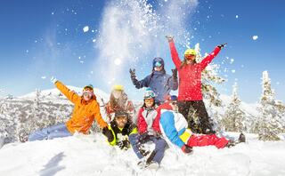 A group of seven cheerful people in colorful winter gear joyfully playing in the snow, throwing snowflakes into the air against a clear blue sky.