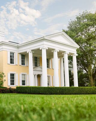The stately yellow Oglebay mansion features grand white columns, lush greenery, and a clear blue sky, exuding elegance and classic architecture.