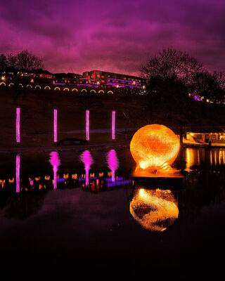 A glowing orb reflects Schenk Lake, illuminated by vibrant purple hues and framed by colorful lights and trees.