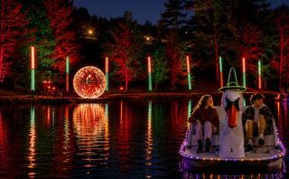 Two people relax on a swan-shaped pedal boat, surrounded by colorful lights reflecting on the water, with a lighted ball in the background.