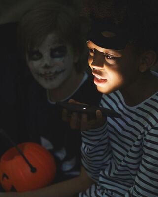 A child with a phone illuminates their face while another in costume watches intently, creating a spooky Halloween atmosphere.