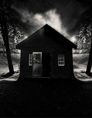 A dark, eerie cabin stands under a smoky night sky, illuminated by distant lights and surrounded by shadowy trees.