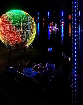A vibrant, illuminated sphere glows on the water, while people gather nearby and boats with colorful lights glide across the dark surface.