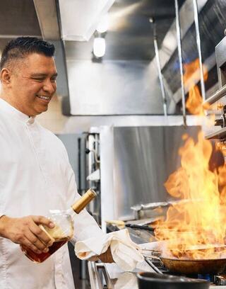 Oglebay chef in a professional kitchen joyfully adds a liquid to a sizzling pan, creating a dramatic burst of flames.