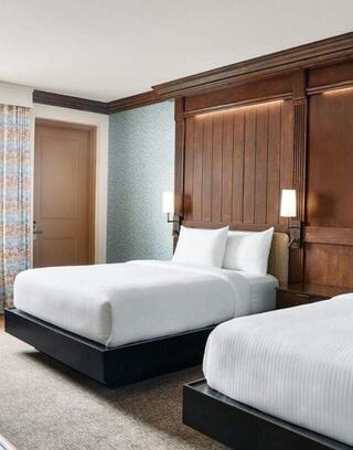 A cozy Oglebay Lodge room features two modern beds with white linens, elegant wood accents, soft lighting, and patterned curtains.