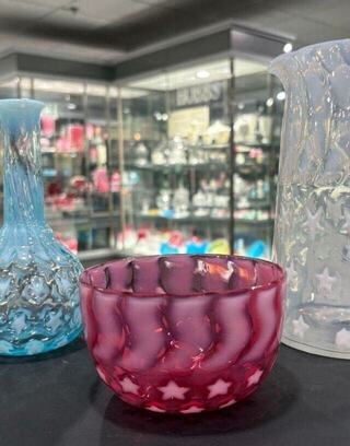 A vibrant collection of glassware featuring a bluish vase, a star-patterned red bowl, and a clear pitcher, all showcased in a retail environment.