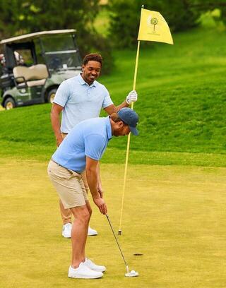 Two men enjoy a sunny day on the golf course, one putting while the other holds the pin, both in casual golf attire.