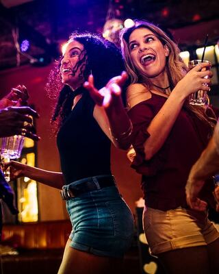 Two women dance joyfully in a lively setting, holding drinks and enjoying the vibrant atmosphere with bright lights and smiles.