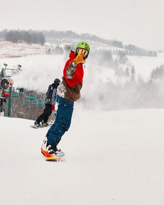 A snowboarder in a bright jacket and helmet gives a thumbs up, with snowy slopes and a ski lift in the background, capturing a fun winter moment.