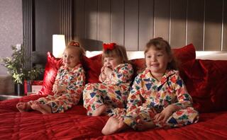 Three smiling girls in colorful pajamas sit together on a red bed, sharing a cozy moment with playful expressions.