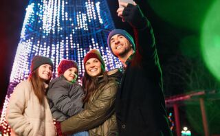 A cheerful family takes a selfie in front of a brightly lit holiday tree, wearing cozy hats and enjoying the festive atmosphere.