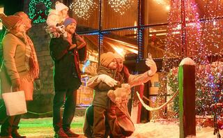 A cheerful family takes a festive selfie near colorful lights and decorations, with snow on the ground, creating a joyful holiday scene.