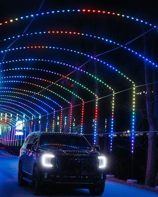 A car drives through a colorful light tunnel, illuminated by vibrant strings of lights in various hues against a dark background.