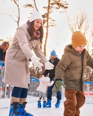 A joyful woman in a beige coat encourages a young boy in a brown hat as they skate on syntheitc ice, surrounded by family and a winter setting.