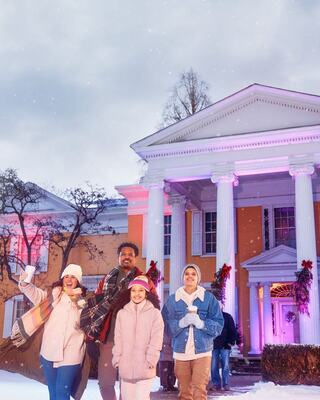 A cheerful group of four stands outside a beautifully decorated house, surrounded by snow and festive lights, celebrating together.