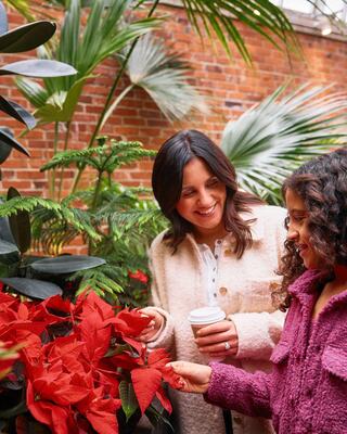 A woman and a girl smile while admiring vibrant red flowers in a lush greenhouse filled with tropical plants.