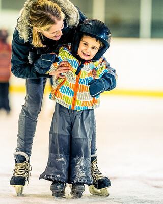 A smiling child in a colorful jacket skates on ice, supported by an attentive adult, both wearing helmets and enjoying a joyful moment together.