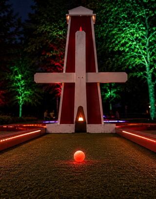 A glowing orange ball sits on green turf, framed by vibrant lights and a large, striking windmill at night.