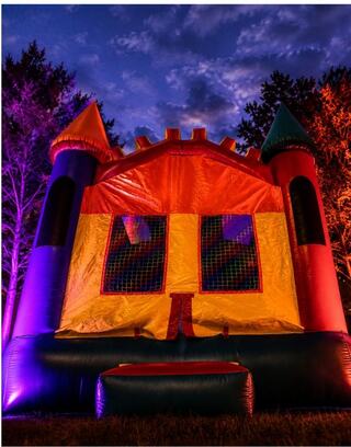 A bounce house glows under a twilight sky, with colorful turrets and illuminated sides, set in an outdoor setting.