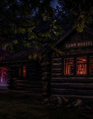 A rustic log cabin with warm lights glowing from the windows, set in a mysterious, foggy forest at night, evokes an eerie atmosphere.