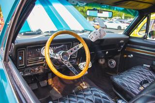 A classic car interior features a wooden steering wheel, black quilted seats, and chrome accents, with an open hood revealing vibrant stripes.