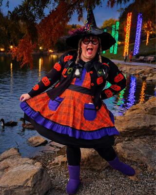 A cheerful person dressed in a whimsical witch costume, with bright colors and fun accessories, poses playfully by a scenic waterfront at dusk.