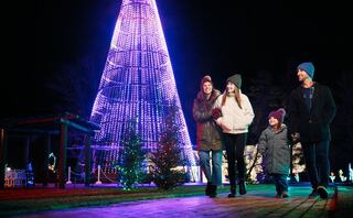 A family enjoys a festive evening walk under colorful Christmas lights, smiling and bundled up against the cold.