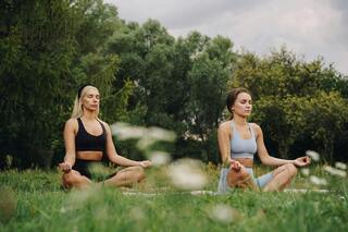 Two women practice meditation outdoors, seated on mats in a peaceful green setting, surrounded by nature and softly swaying grass.