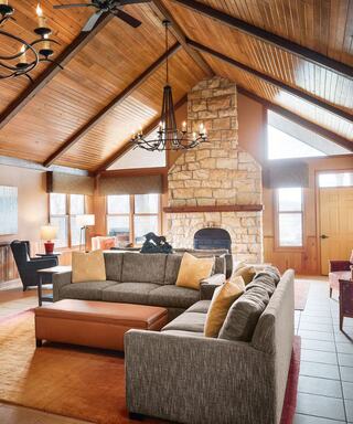 A cozy living room features a large sectional sofa, stone fireplace, wooden beams, and ample natural light from expansive windows.