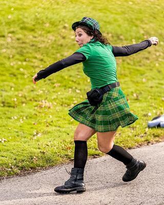 A woman in a green shirt and plaid skirt dances energetically, wearing knee-high socks and boots, enjoying a festive atmosphere outdoors.