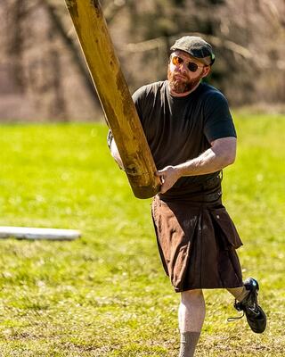 A man in a kilt and sunglasses lifts a wooden beam in a grassy field, showcasing strength and determination.