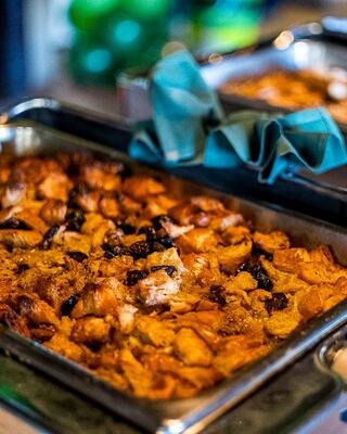 Warm, golden-brown bread pudding topped with dark raisins, served in a metal tray with green napkins, ready for a delicious treat.