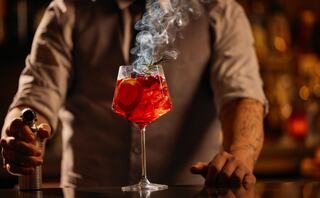 A vibrant cocktail, garnished with citrus, emits a smoky vapor, while a bartender expertly prepares it against a blurred, colorful backdrop.