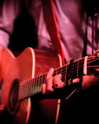 A close-up of a hand strumming an acoustic guitar, showcasing the instrument's wooden body and a capo on the neck, with soft lighting.